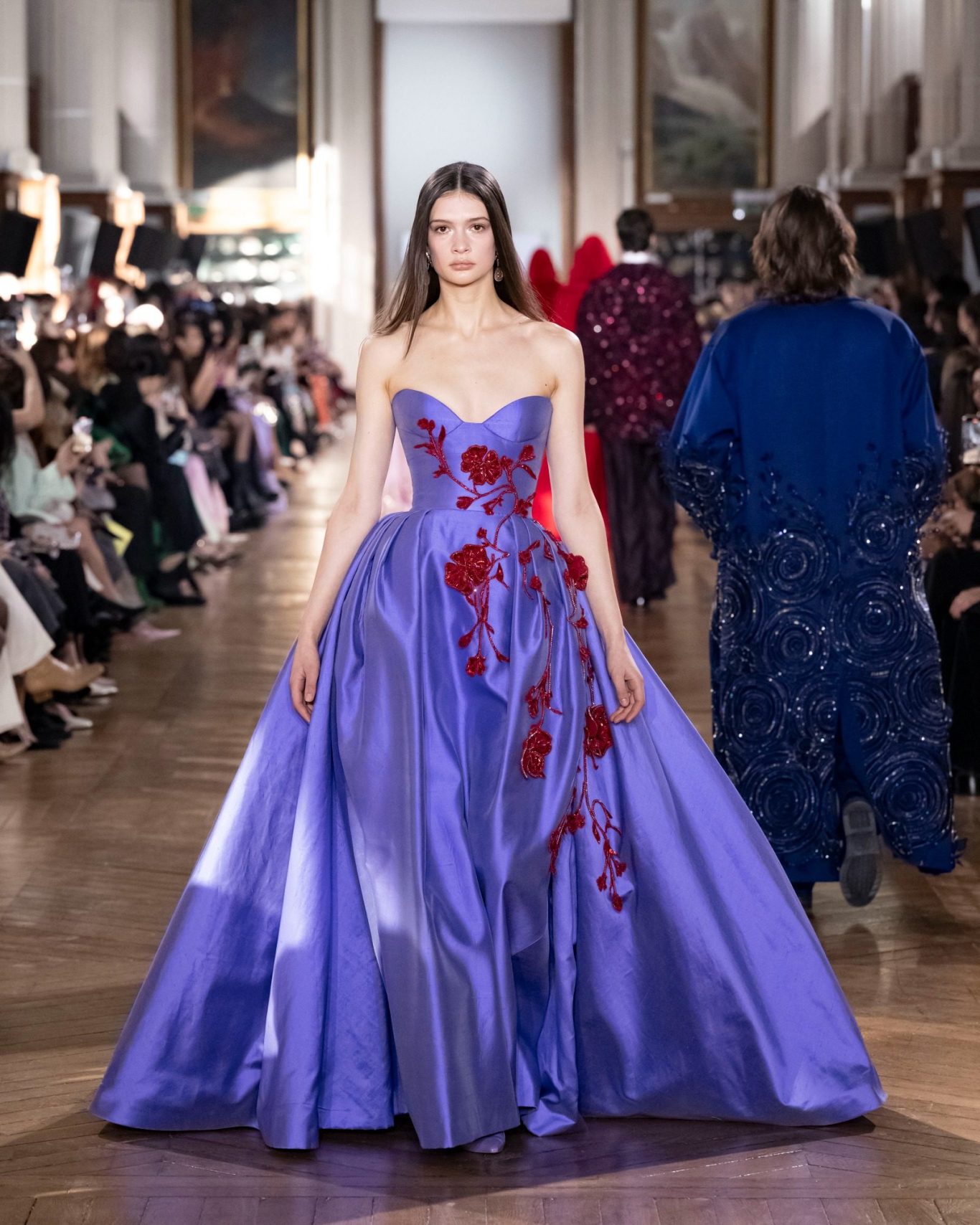 A woman in a metallic embellished Zuhair Murad evening gown with thin straps stands against a deep red background, exuding elegance.