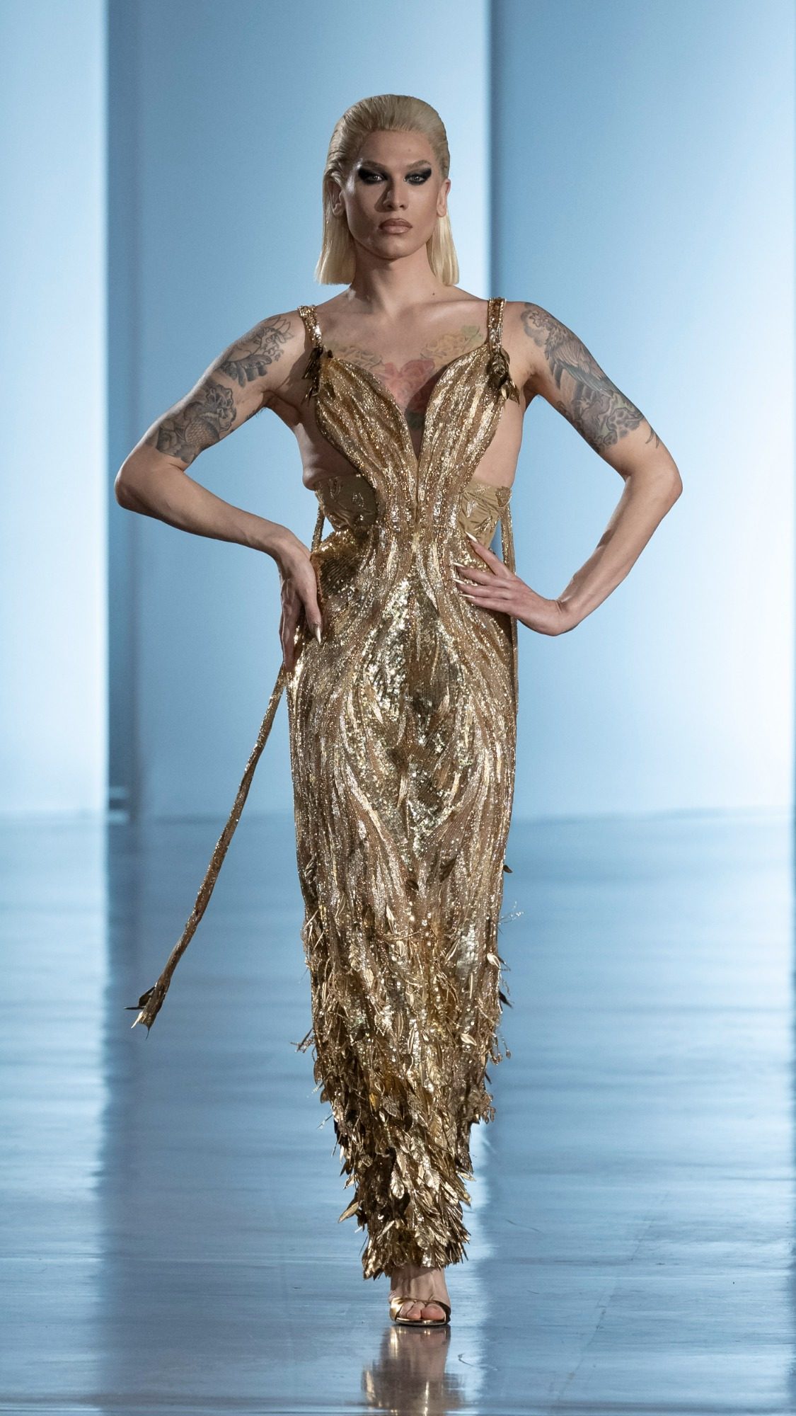 A woman in a metallic embellished Zuhair Murad evening gown with thin straps stands against a deep red background, exuding elegance.