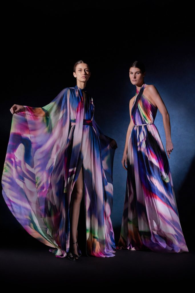 Two women wearing vibrant, flowing Zuhair Murad gowns with abstract multicolor patterns pose against a dark background.