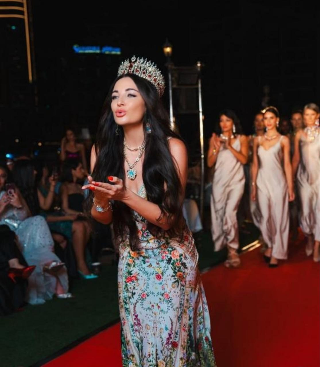 A woman in a metallic embellished Zuhair Murad evening gown with thin straps stands against a deep red background, exuding elegance.