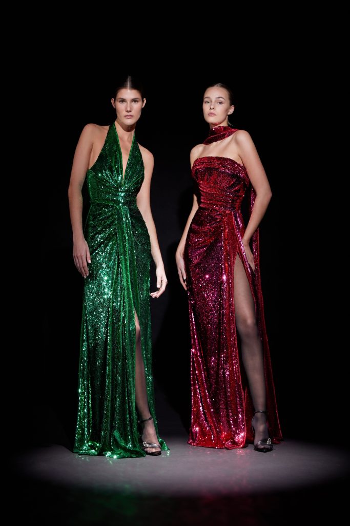 Two women pose in dazzling Zuhair Murad gowns: one in emerald green and the other in ruby red, both with high slits.