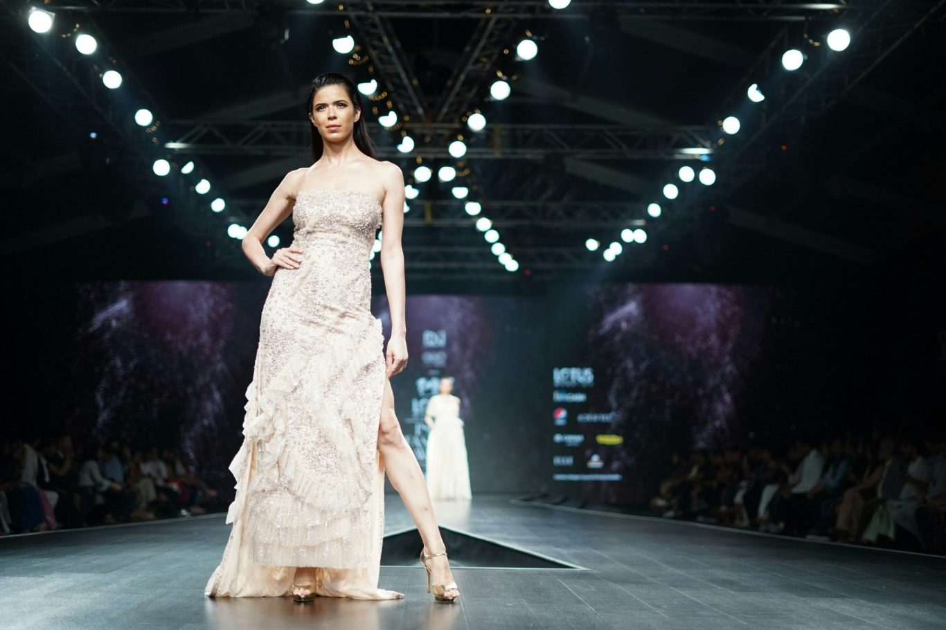 A woman in a metallic embellished Zuhair Murad evening gown with thin straps stands against a deep red background, exuding elegance.