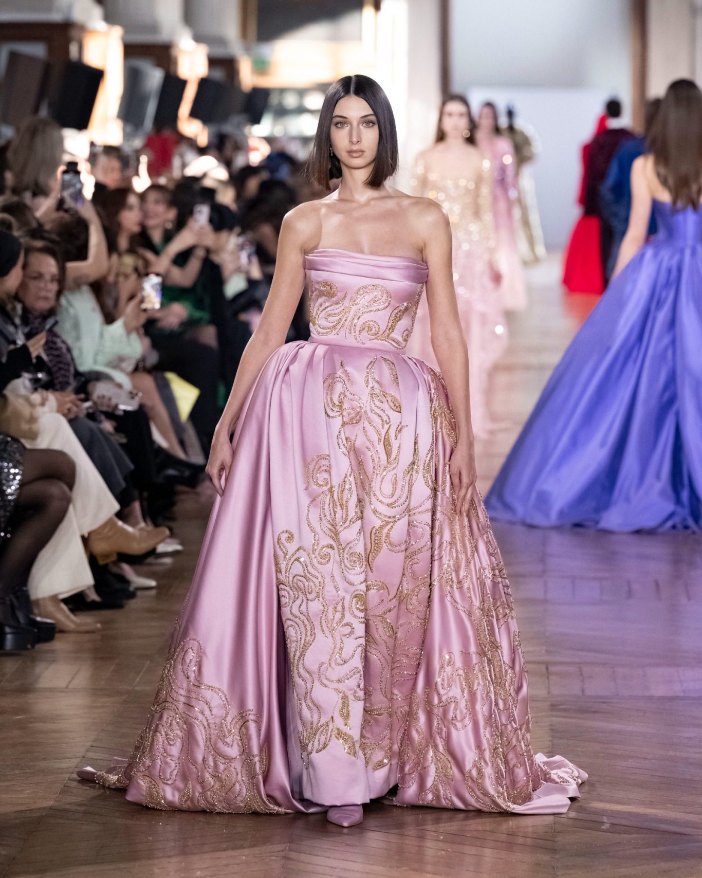 A woman in a metallic embellished Zuhair Murad evening gown with thin straps stands against a deep red background, exuding elegance.